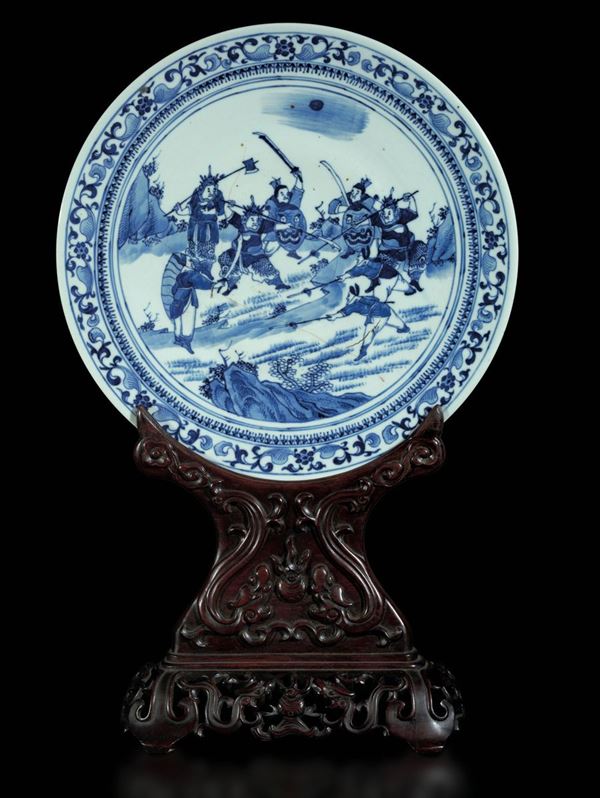 Piatto in porcellana bianca e blu raffigurante scena di battaglia e decori floreali su stand in legno di Homu, Cina, Dinastia Qing, epoca Guangxu (1875-1908)