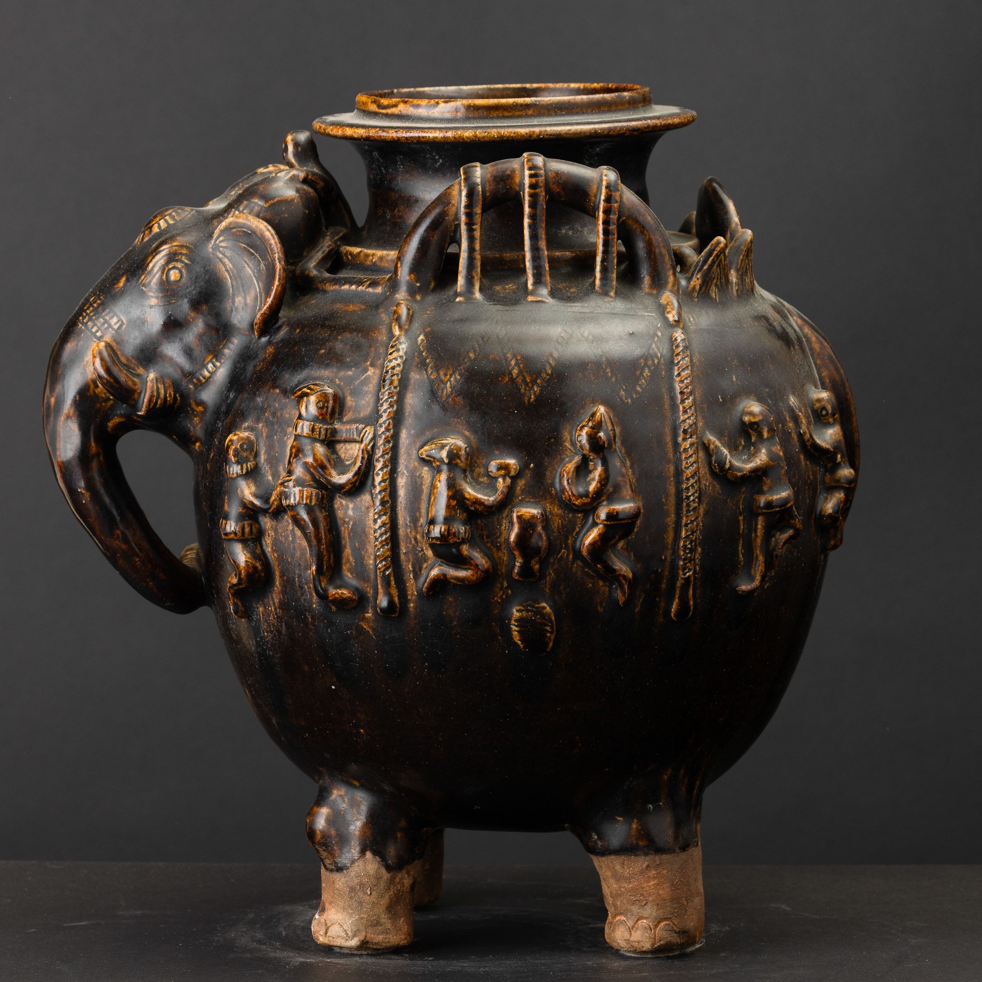Arte Khmer, Vaso in terracotta invetriata a fondo marrone con testa di elefante e figure a riliev...
